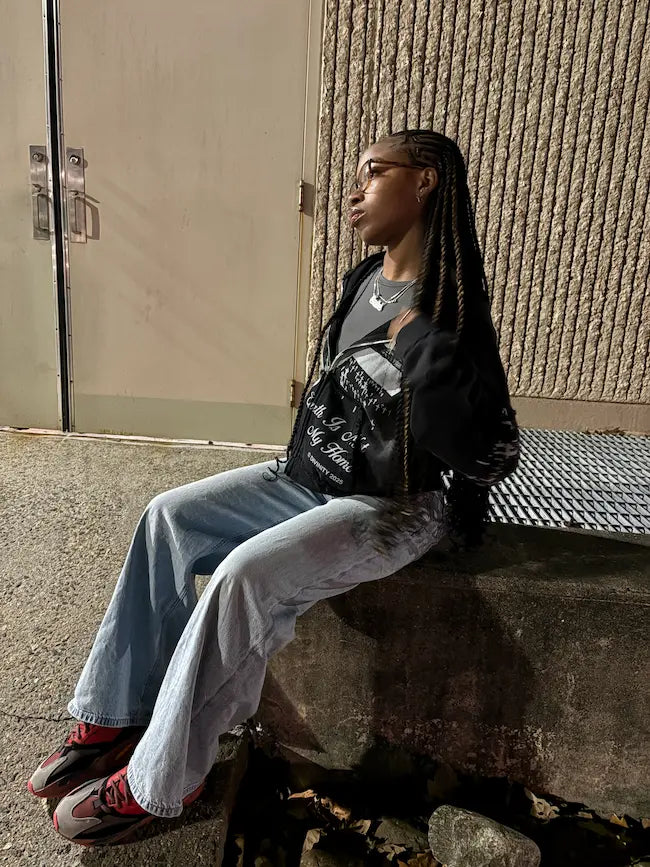 Person sitting on a ledge wearing a black jacket and light blue jeans.